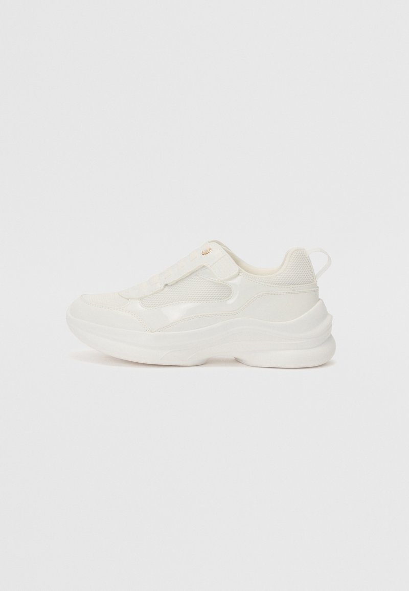 Baskets blanches épaisses avec dessus en mesh et cuir, semelle épaisse et languette de traction au talon, présentées de profil sur un fond blanc.