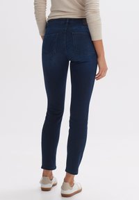 Dunkelblaue Skinny-Jeans mit einer glatten Denim-Oberfläche, ausgestattet mit zwei Gesäßtaschen und einer mittelhohen Taille. Getragen mit weißen Sneakers.