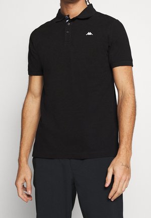 Mężczyzna ubrany w czarną, krótkorękawową koszulkę polo z białym logo na lewej piersi oraz czarne spodnie na białym tle.