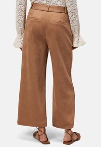 Pantalons larges en tissu marron avec une texture douce, dotés d'une ceinture, de poches arrière et d'un ourlet propre. Portés avec des sandales plates marron.