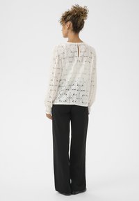 Kiharate ja, kuldkettide kõrvaehteid kandva, valge lillemustriga pitsbluusiga ja mustade laia jala pükstega naine, seljaga kaamerale, valgel taustal.