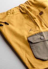 Des shorts en tissu jaune présentent une taille élastique, un cordon de serrage avec des boutons en plastique noir, une poche grise avec un bouton et des coutures décoratives.