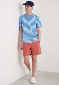 Randig blå och vit kortärmad skjorta matchad med korallfärgade shorts; vita sneakers. Avslappnad outfit mot en neutral väggbakgrund.