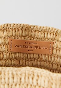 Bolso de tote de paja trenzada con una etiqueta de cuero marrón claro que dice "LE CABAS VANESSA BRUNO PARIS". Presenta tonos naturales y un acabado texturizado.