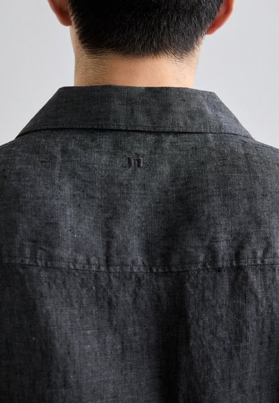 Schwarzes Leinenhemd mit einem umschlagbaren Kragen, sichtbarer Textur und einem kleinen bestickten Logo am Nacken. Einfaches, minimalistisches Design.