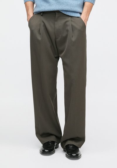 Széles lábú nadrág sötét olíva zöld anyagból, elülső redőkkel, oldalsó zsebekkel és sima textúrával, fekete cipőkkel párosítva.