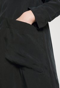 Svart tygplagg med en synlig sidoficka, utrustad med en mjuk yta och en subtil glans, som visar upp rena sömmar och en avslappnad design.