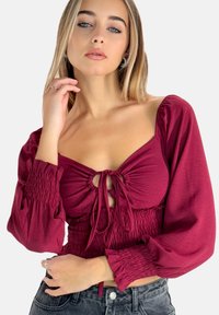 Bordeaux crop-top met puff-mouwen, een gesmokte buste en een strikdetail aan de voorkant. Gemaakt van een zachte stof met een gestructureerde afwerking.