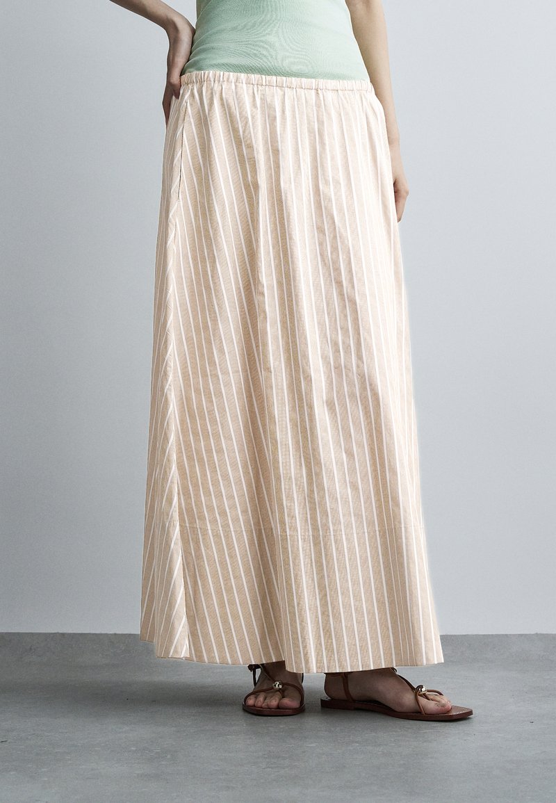 Woman wearing a beige and white vertically striped maxi skirt with an elastic waistband and brown flat sandals on gray floor.