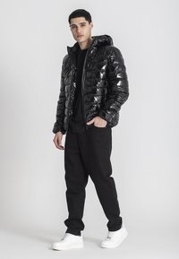 Giacca a vento nera con texture brillante e trapuntata, con cappuccio, indossata sopra una t-shirt nera e abbinata a pantaloni neri larghi e sneakers bianche.