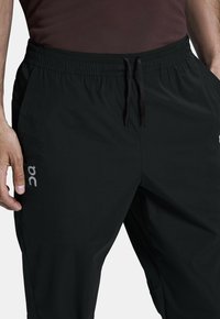 Pantalons de sport noirs en tissu léger, avec une taille élastique dotée d'un cordon de serrage et des poches latérales. Affiche un logo gris.