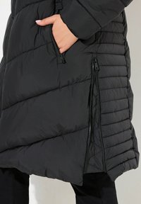 Svart pufferjacka med horisontellt quiltat mönster, sidofickor med dragkedja och en slät textur. Har en avslappnad passform och justerbara öppningar.