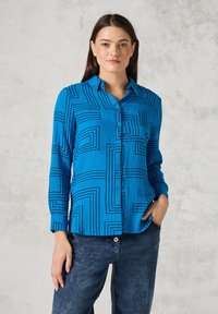 Camisa azul de botones hecha de tejido texturizado con un patrón geométrico de líneas negras. Mangas largas y detalle de bolsillo en el pecho.