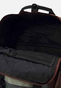 Hnědý a zelený látkový batoh má uvnitř černou podšívku, zipovou kapsu a texturovaný klopu se suchým zipem.