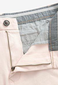 Pantalones rosa claro con cremallera en la parte delantera, cuentan con un forro interior a rayas azul oscuro y un botón negro. Detalles de costura a contraste visibles.
