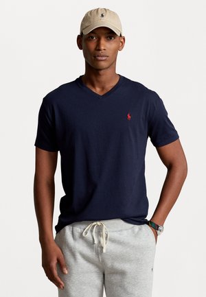 T-shirt a V-collo blu della marina con logo rosso sul petto, abbinato a pantaloni della tuta grigi. La figura indossa un cappellino beige, mostrando un outfit informale.
