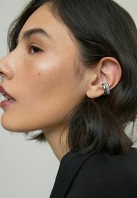 Sølvørering med en struktureret design, der har skimrende accenter. Cirkelformet, båret på øret af en person med mørkt hår.