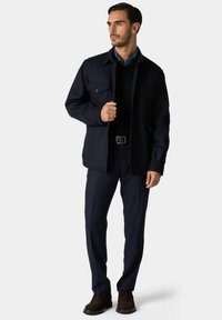 Marineblauw overhemdjack met twee borstzakken, gedragen over een zwarte trui, gecombineerd met marineblauwe broek en bruine schoenen.