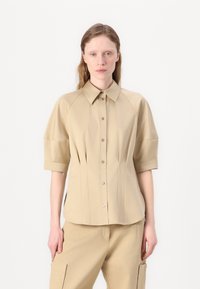 Camicia beige a maniche corte con chiusura a bottoni, colletto a punta e design plissettato. Realizzata in tessuto liscio con una vestibilità strutturata.