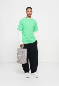 Tricou verde din bumbac peste o cămașă albă cu mâneci lungi, asociat cu pantaloni negri. Geantă gri tip tote cu logo-ul Nike, adidași albi, fundal neutru.