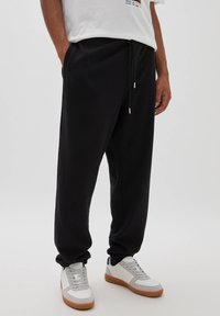 Svarta joggingsbyxor med elastiskt midjeband och snörning. Har sidofickor och avsmalnande muddar. Matchas med grå och vita sneakers.