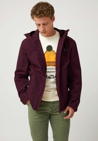 Chaqueta impermeable color burdeos con capucha, cierre con cremallera y puños elásticos, llevada sobre un suéter crema que presenta un diseño gráfico en naranja y negro.