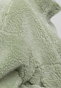 Uldne grøn fleece-stof med en tekstureret overflade, der har lynlås lukning. Materialet er blødt og plyssigt, ideelt til varme.