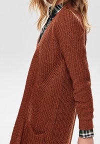 Vrouw in een roestkleurige gebreide cardigan over een groen-wit geruit shirt, zijaanzicht tegen een effen achtergrond.