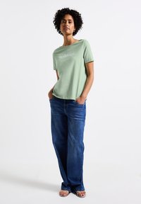 T-shirt en coton vert clair à manches courtes, présentant un motif imprimé. Associé à un jean en denim bleu ample et des sandales.