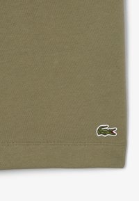 Camiseta de algodón verde oliva con una textura suave y un pequeño logo de cocodrilo en blanco y verde bordado en el dobladillo inferior derecho.