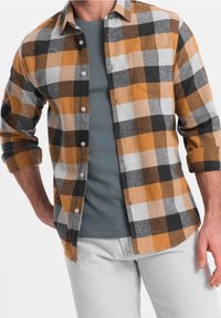 Koszula flanelowa w pomarańczowo-szaro-czarną kratę. Posiada zapięcie na guziki z przodu oraz jedną kieszeń na piersi. Noszona na szarym t-shircie.
