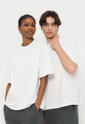 Dvaja mladí dospelí v bielych nadrozmerných tričkách a tmavosivých nohaviciach stoja tesne vedľa seba na jednoduchom bielom pozadí.