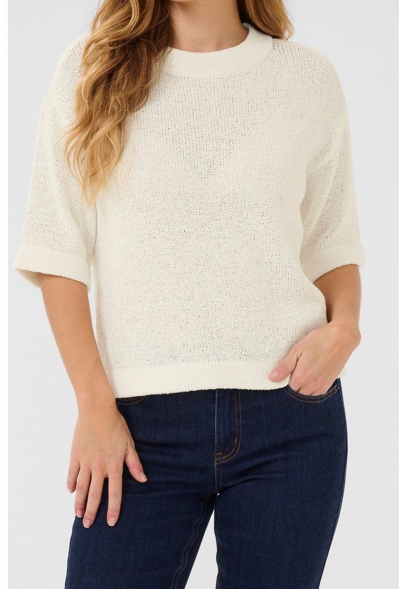 Pull blanc en maille avec manches courtes, col et ourlet côtelés, offrant une coupe ample et une texture légère. Associé à un jean en denim foncé.