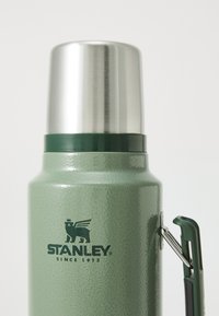 Green, textured thermos with a shiny stainless steel lid. Features a handle and black logo with a bear and crown. Sturdy design.