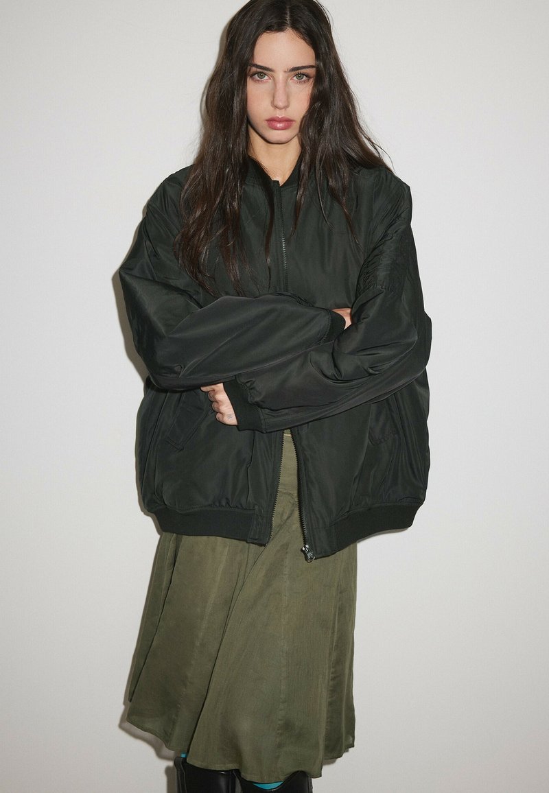 Femme aux longs cheveux foncés portant une veste noire surdimensionnée par-dessus une jupe vert olive sur un fond blanc uni.
