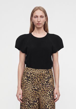 Femme aux longs cheveux blonds portant un haut noir avec des épaules rembourrées et un pantalon taille haute imprimé léopard, debout devant un fond uni.
