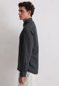 Dunkelgraues Hemd mit knöpfbarem Kragen und langen Ärmeln, das eine subtile Textur und scharfe Manschetten aufweist. Getragen über hellgefärbten Hosen.