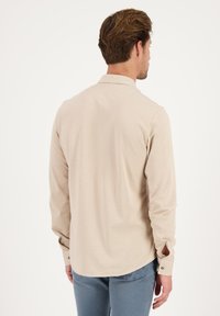 Camisa de manga larga beige con cuello, que presenta botones en los puños y un detalle de abertura. Elaborada con un tejido suave de textura lisa.