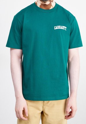 Hombre con camiseta verde Carhartt y pantalones beige, de pie frente a un fondo claro y liso.