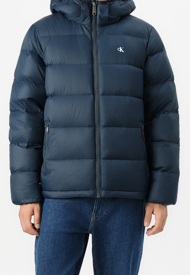 Doudoune bleu marine avec fermeture éclair à l'avant, poches latérales zippées, capuche et petit logo blanc "cK" sur la poitrine gauche, portée avec un jean bleu.