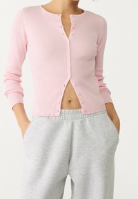 Roze geribbelde cardigan met lange mouwen en een ronde halslijn. Voorzien van knopen aan de voorkant en een cropped ontwerp, gecombineerd met grijze joggingbroek.