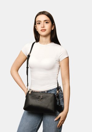Junge Frau trägt ein weißes, eng anliegendes T-Shirt und blaue Jeans, hält eine schwarze Lederschultertasche mit goldfarbenen Beschlägen vor hellem Hintergrund.