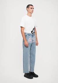 Camiseta blanca de manga corta con un gráfico de dibujo animado negro en la parte frontal, combinada con unos jeans de cintura alta de color azul claro con un diseño rosa. Zapatillas negras.