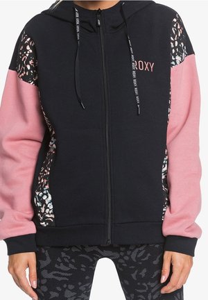 Frau trägt einen schwarzen Kapuzenpullover mit Reißverschluss, pinken Ärmeln und abstrakt gemusterten Einsätzen, kombiniert mit schwarzen gemusterten Leggings.