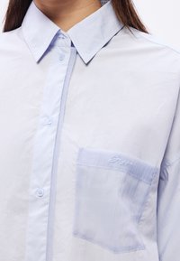 Chemise bleu clair à boutons avec col pointu et poche poitrine, portée par une personne aux longs cheveux foncés visibles au niveau du cou.