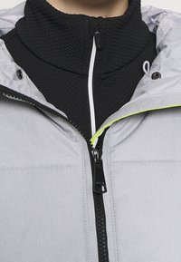 Gros plan d'une personne portant un vêtement zippé noir texturé sous une veste gris clair avec une fermeture éclair et des boutons-pression noirs.