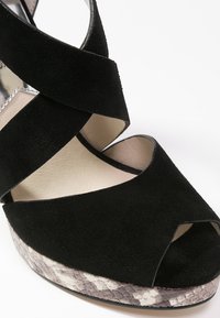 Zwarte nubuck hoge hakken sandaal met gekruiste banden en een open teen. Textuur met slangenprint op het plateau biedt contrast langs de zool.