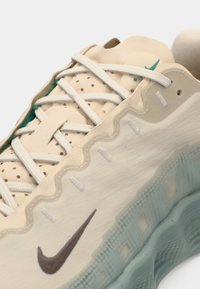 Nike sneakers har en cremefarvet overdel med tekstureret mønster og perforeringer, hvide snørebånd og en lys teal gummisål med et bølget design.