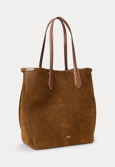 Sac fourre-tout en daim marron avec deux longues poignées en cuir et un petit logo doré près du bas au centre sur un fond clair uni.