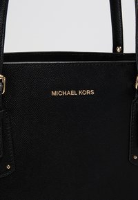 Sac à main texturé noir avec logo "MICHAEL KORS" doré, double anses robustes et accessoires métalliques discrets, offrant un design sophistiqué.